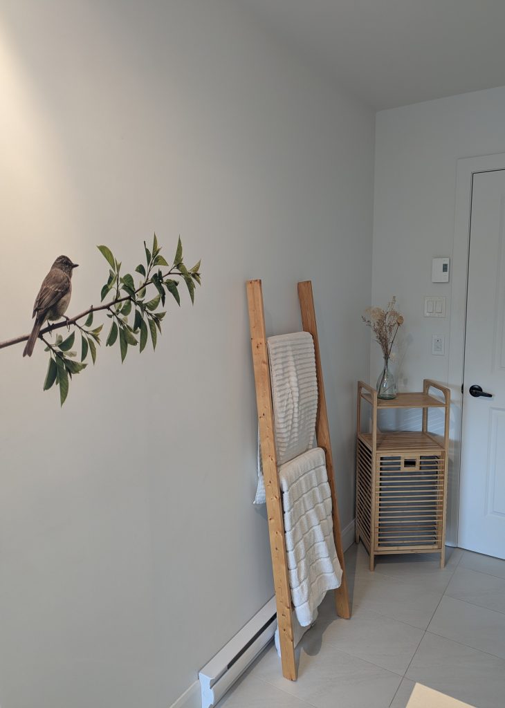 Impression murale réaliste d'un oiseau sur une branche verte, ajoutée sur le mur blanc d'une salle de bain de style zen.